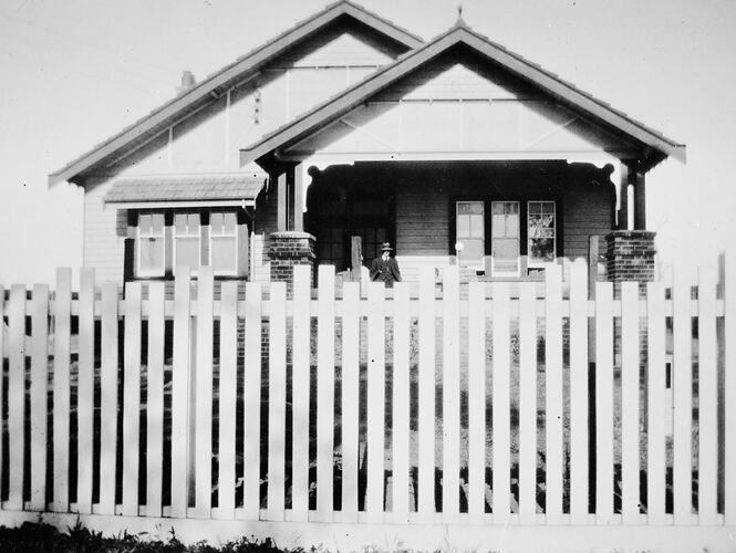 Man standing on the verandah of a newly built house. There is a picket fence in the foreground.