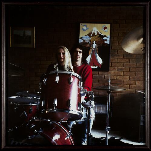 Woman and man sit behind red drum kit. Brown brick wall with picture behind them.