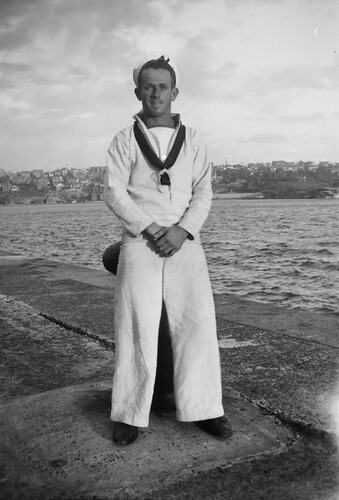 Man in sailors uniform leaning against mooring bollard next to water.