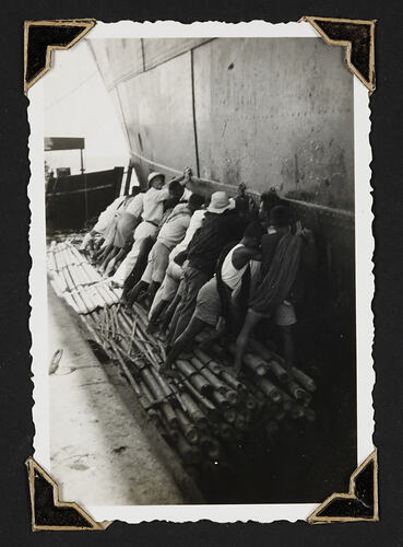 Row of men standing on pile of bamboo pushing the side of a ship.