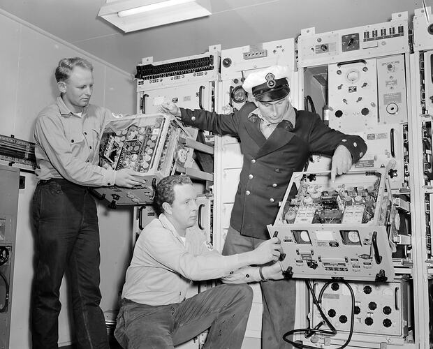 Three men holding two large computer parts in front of a large computer floor to ceiling computer.