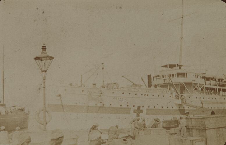 Ship with red cross on side berthed at dock busy with workers and stacks of cargo.