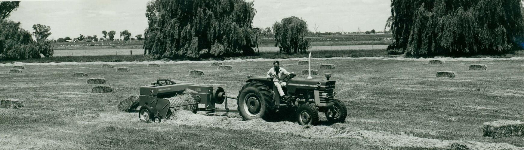 A man driving a tractor pulling a baler in a field with willow trees in background.