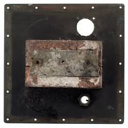 Black metal square (burnt) with holes around edges and two larger ones within. Grey metal rectangular in centre.