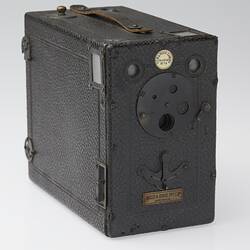 Black box-shaped camera with round metal plate and badges. Three quarter view.