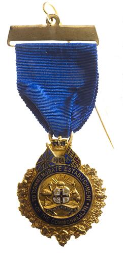 Gold medal with wreath border and blue enamel. Suspended from blue ribbon.