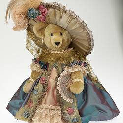 Light brown plush bear wearing lavish cream hat with feather and ornate dusty pink dress and blue grey jacket.