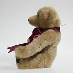 Profile of seated light brown plush bear with red ribbon around neck.