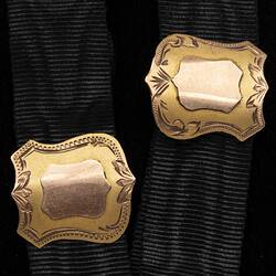Detail of two black ribbed straps, each with a central gold shield-shaped sliding attachment.