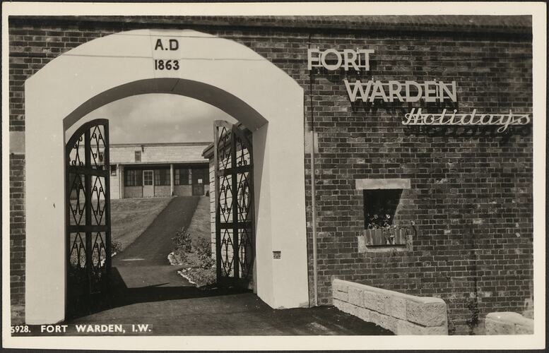 White arched entrance through a brick wall. Wrought iron gates and sign to right on wall. Building visibile through entrance.
