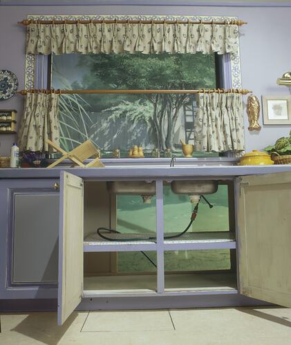 Kitchen studio set, open pale blue cupboards below metal sink show plumbing.
