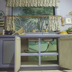Kitchen studio set, open pale blue cupboards below metal sink show plumbing.