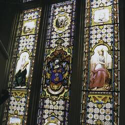 Three tall thin stained glass window panels. Women each side, coat of arms in centre.