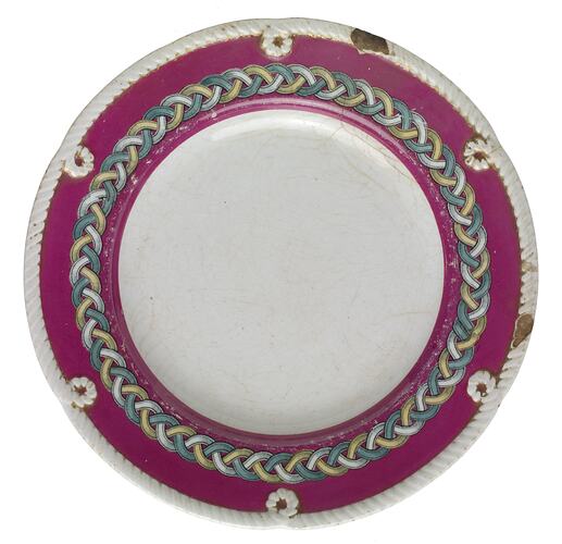 White dinner plate with raised cable edging. A deep pink border features a gold, green and white woven cable d