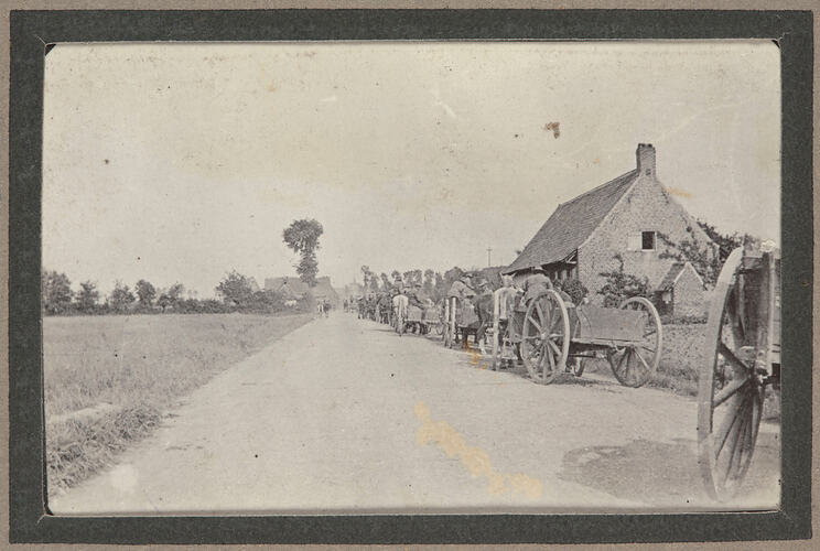 Convoy of men and horse drawn cartridges on a country road, with houses and trees.