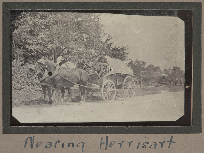 Two horses pulling a large, covered cart along a tree lined, dirt road, truck with servicemen in background.