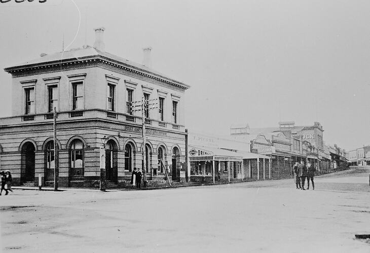 Street view with two storey building at left on corner. Dirt road and few figures stand at right.