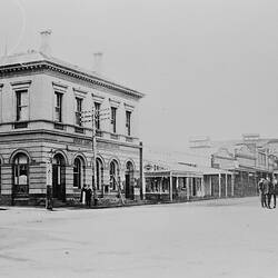 Street view with two storey building at left on corner. Dirt road and few figures stand at right.