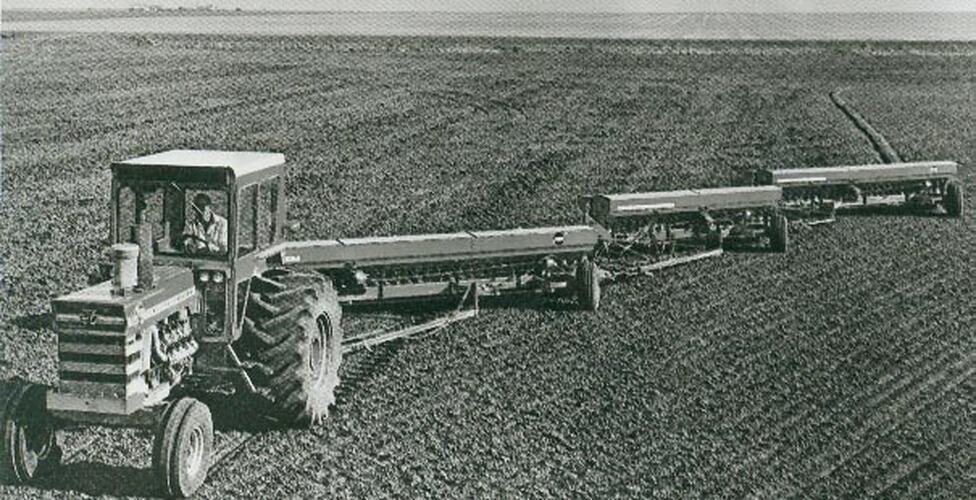 Man driving a tractor across a field with 3 seeders hitched in a line behind.