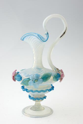 Blue blown-glass ewer, spiral fluted neck, serpentine handle, flattened bulbous body layered with pink, blue green flowers and leaves.