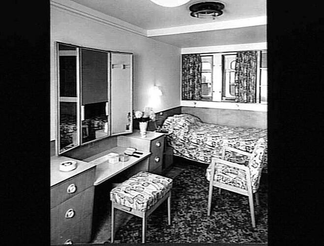 Ship interior. Single bed against wall. Dressing table and stool at left. Chair at right.