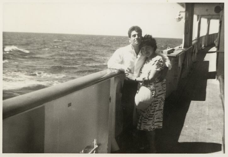Man and woman cuddle and pose on a ship deck with the open ocean behind them.