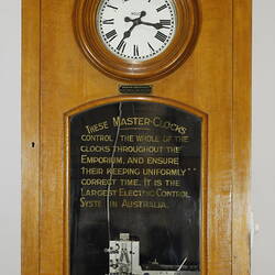 Detail of wooden clock. White clockface with glass panel below that has gold lettering.