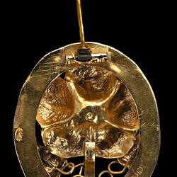 Back of oval decorative gold brooch containing a flower inside a larger flower, all within an oval border entwined with gold ribbon. Pin open.