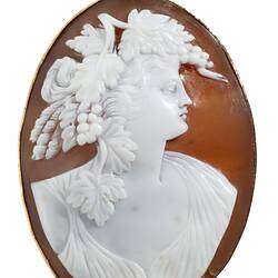 Oval brooch, white cameo of classical female facing right with a headpiece of grapes, leaves and feathers. Brown background set in polished gold border.