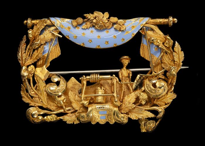 Rectangular gold brooch. Ornate detail, open centre. Blue enamel drapery at top. Miner, tools and foliage.