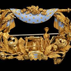 Rectangular gold brooch. Ornate detail, open centre. Blue enamel drapery at top. Miner, tools and foliage.