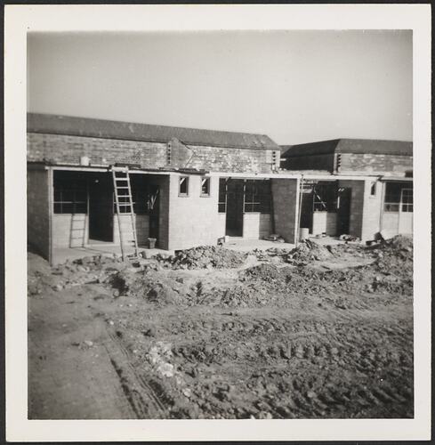 Units in the process of being built. Uneven dirt in foreground and ladder propped against building. No windows or doors.