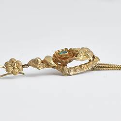 Back of gold pendant earring. Comprised of four connected sections with floral and leaf motifs. Gold fringe hangs from the bottom. Profile.