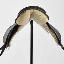 Horse racing saddle. Padded dark brown leather. Double layer beneath saddle flap. Two leather straps on either side. Front view.