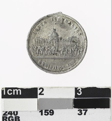 Round silver coloured medal with building, text above and below.