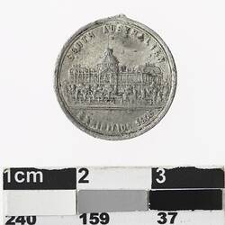 Round silver coloured medal with building, text above and below.