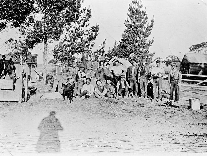 17 men and boys pose in two rows. There are dogs and a horse in the scene. Tent and hut in background.