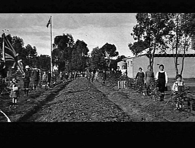 Children lined up on both sides of a road, man holding flag on right, building on left.