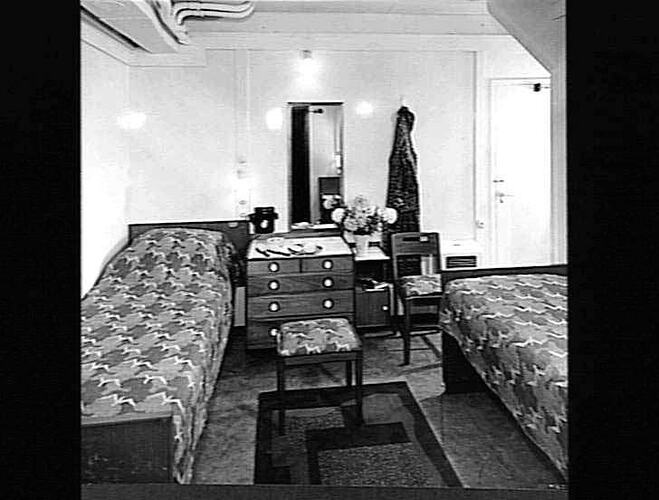 Ship interior. Two single beds, one against wall. Drawers and stool in between. Chair at right.