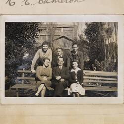 Four men and two women posing in a garden, with a large brick factory building behind them.