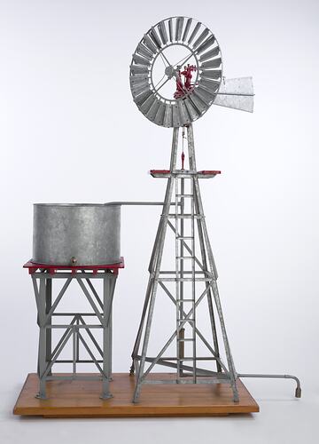 Model water pumping windmill next to water tank on a stand. Vanes facing front.