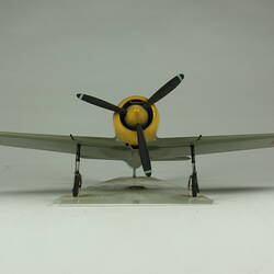 Green grey plane model with red spot on each wing and fusleage. Yellow nose, black propeller. Front view.