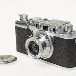 Black camera, metal trim, cap, lens, dials at top corners. Front angle view.