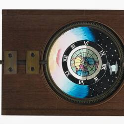 Rectangular wooden framed glass slide with handle at left. Central circular glass image of day and night.