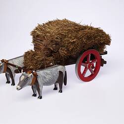 Model of two grey bullocks pulling a hay wagon with haystack and red wheel. Three quarter view.