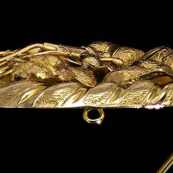 Profile of oval decorative gold brooch containing a flower inside a larger flower, all within an oval border entwined with gold ribbon. Pin open.