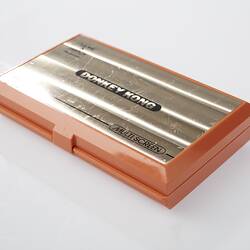 Closed orange plastic, handheld game console. Top has metallic label and text.