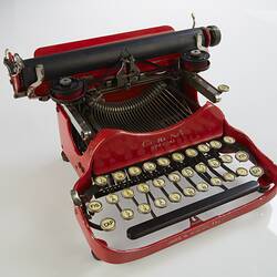 Red metal typewriter, 32 keys, black plastic spacebar at front, black roller for paper at top back. Front.