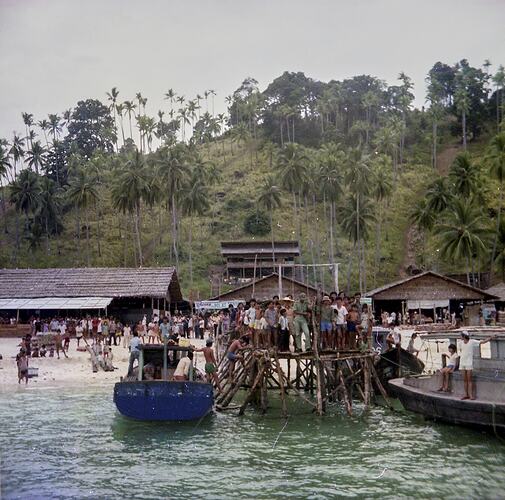 Busy scene of crowds amongst boats and a beach at at Vietnamese Refugee Camp. Heavily treed hill in background.