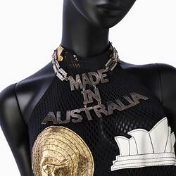 Detail of metal MADE IN AUSTRALIA necklace and top of black halterneck mesh dress with gold and white applique cut outs.